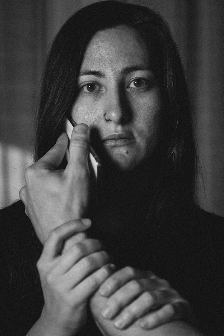 A woman looks forward while holding a phone to her ear. A hand is gripping her wrist firmly. Black and white image.