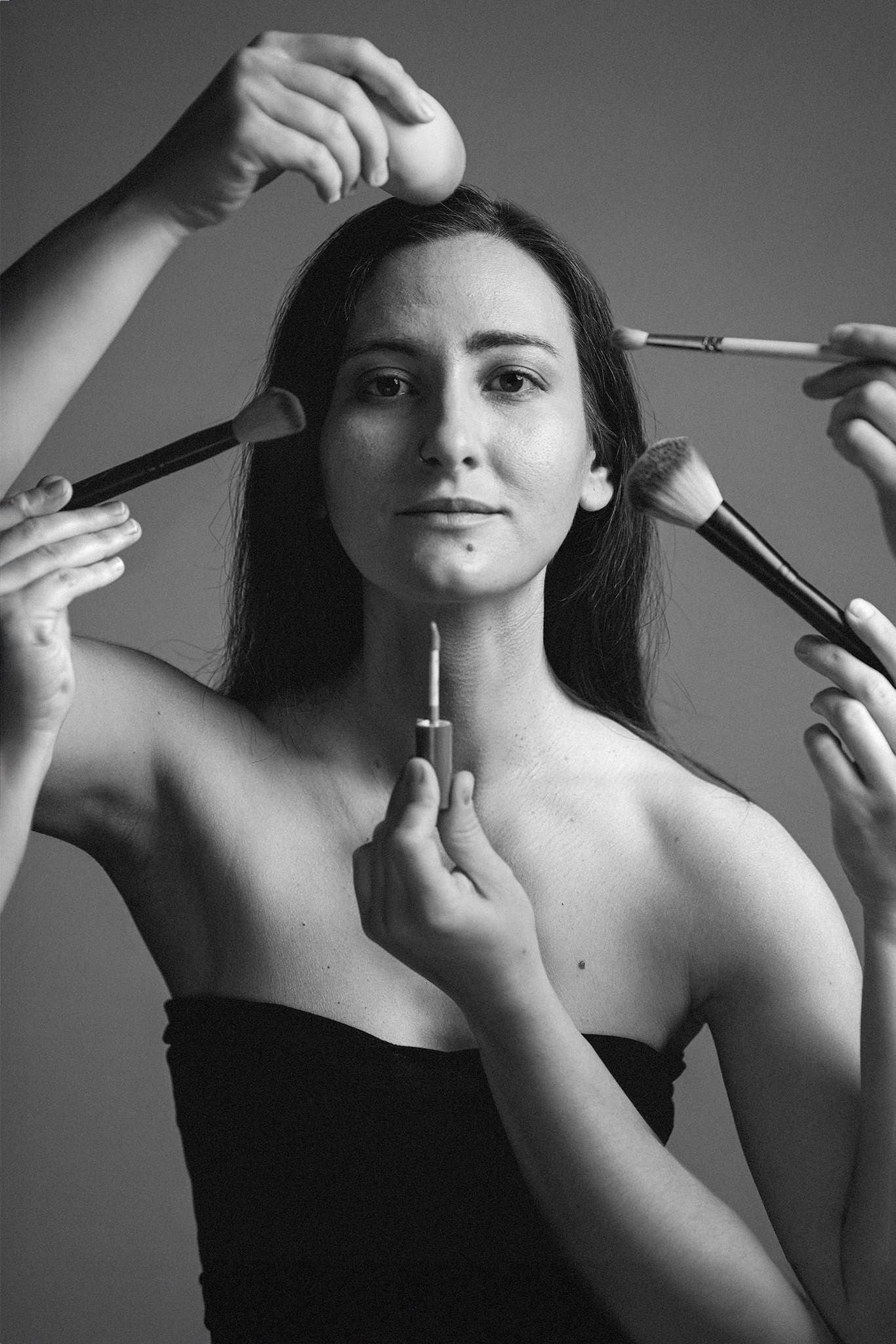 A black-and-white portrait of a woman surrounded by multiple hands holding makeup tools, applying various products to her face. She gazes calmly at the camera.