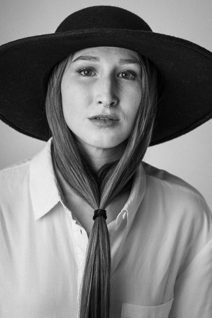 Black and white portrait of a woman wearing a wide-brimmed hat and a nose ring, with her long hair styled uniquely under her chin.