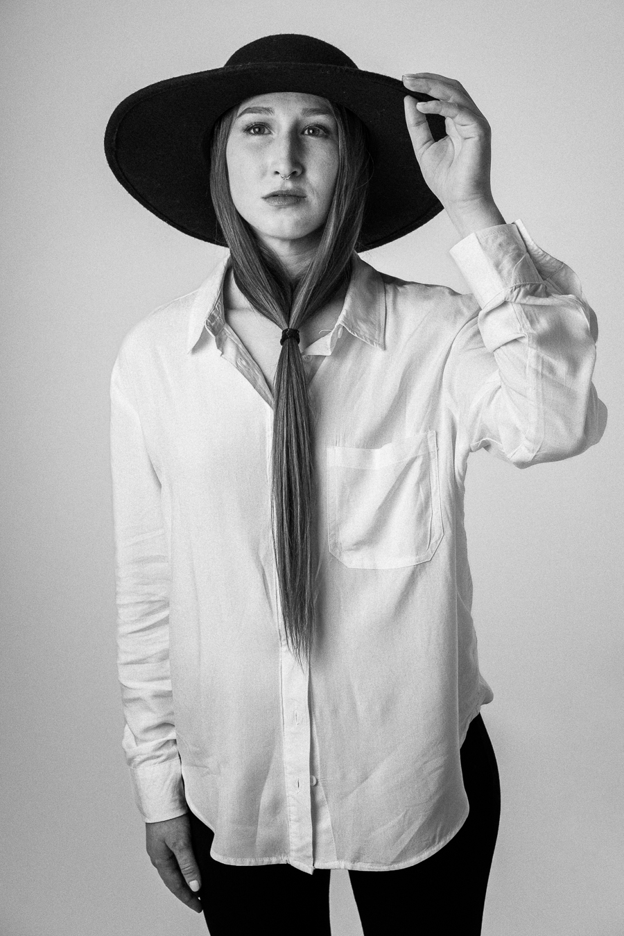 Black and white portrait of a person wearing a large hat and a white shirt, standing in front of a neutral background.
