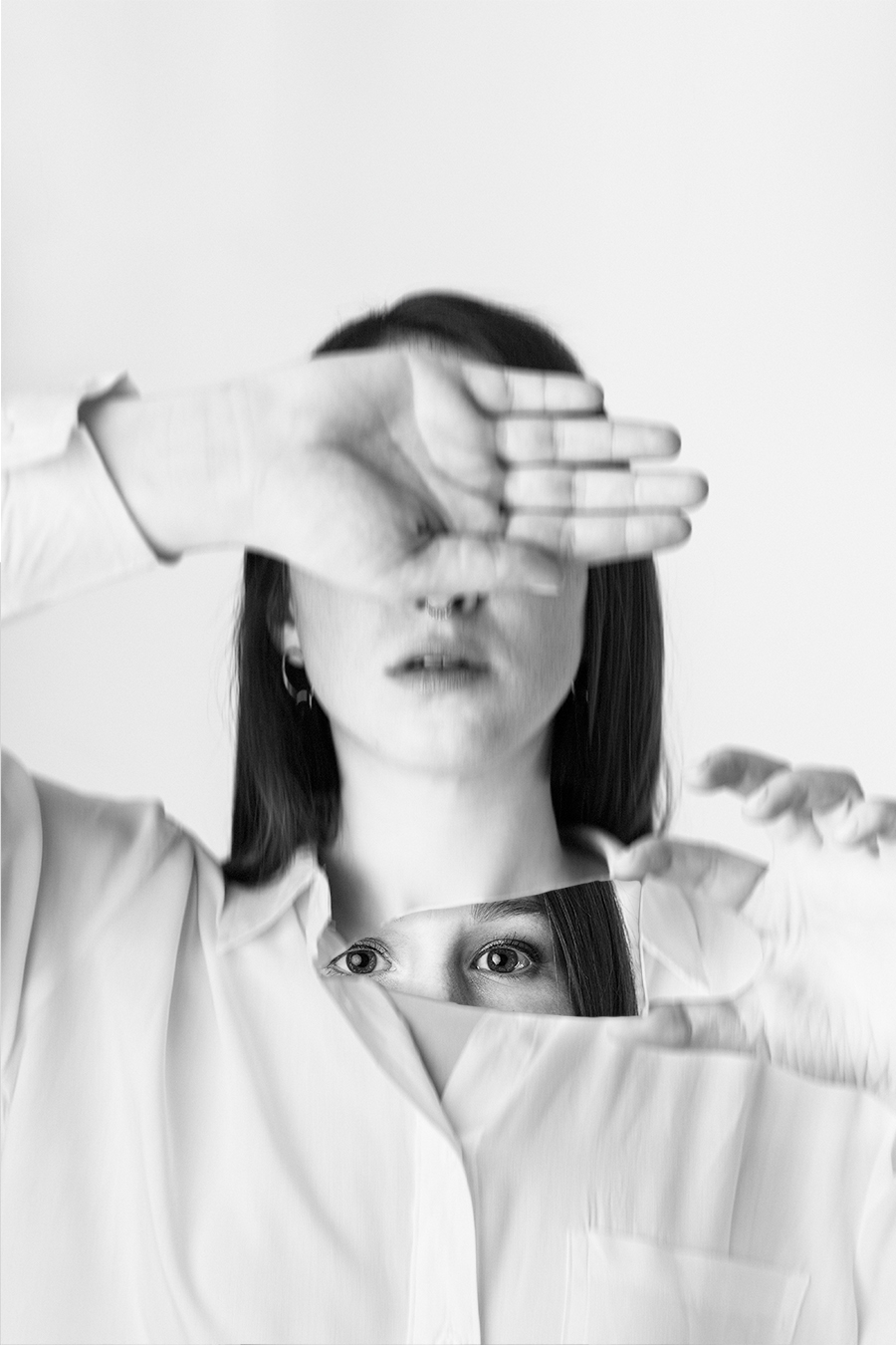Black and white artistic portrait of a woman partially covering her face with her hand, holding a mirror reflecting her eyes, conveying themes of reflection and identity.
