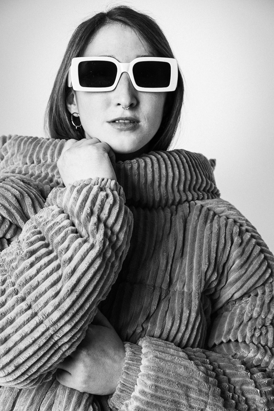 Black and white portrait of a person wearing large white sunglasses and a textured, ribbed coat backwards, showcasing contemporary fashion style.