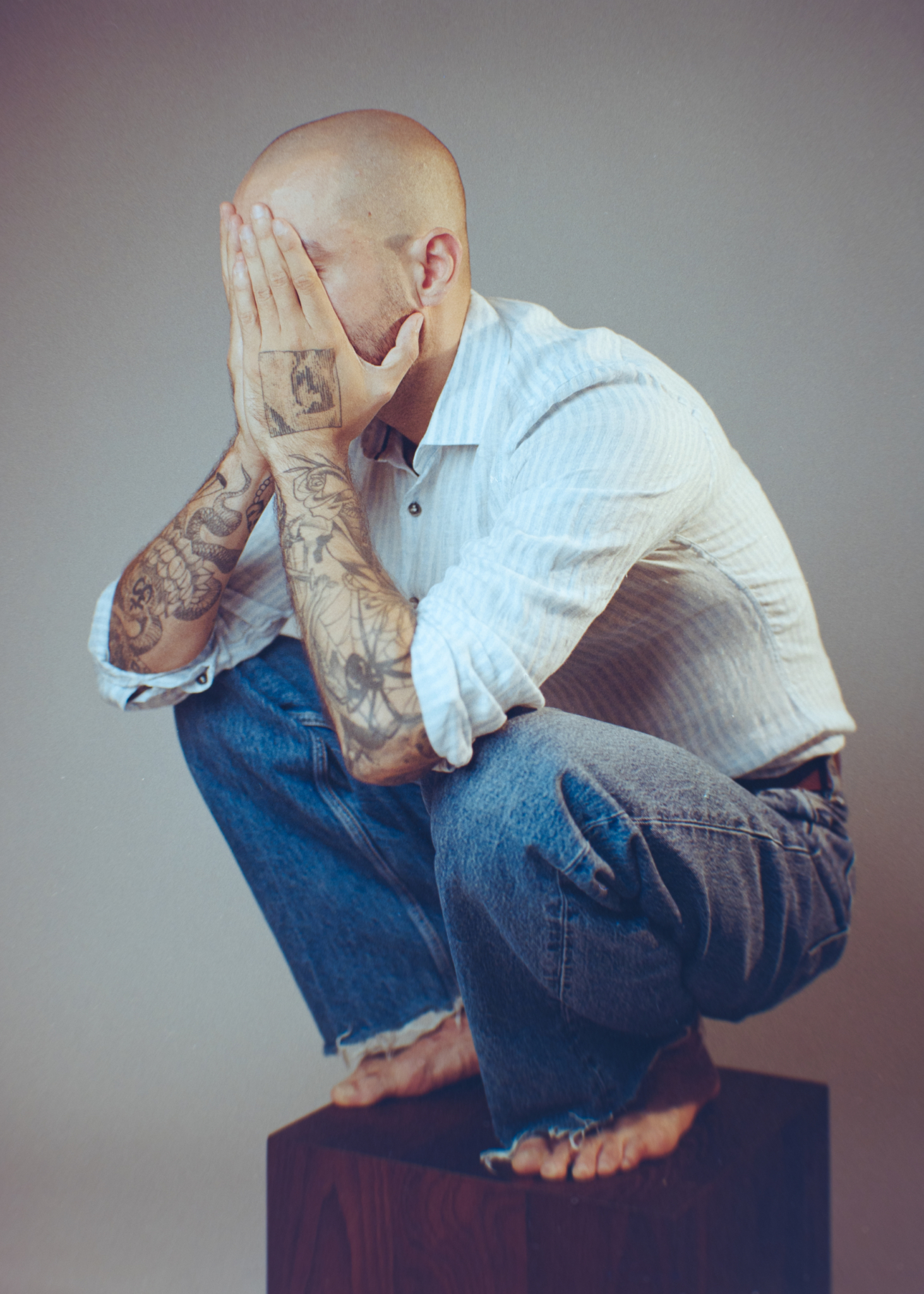 Coloured analogue portrait of a person with tattoos crouching on a wooden block, wearing a striped shirt and jeans, featuring thoughtful expression and artistic pose.