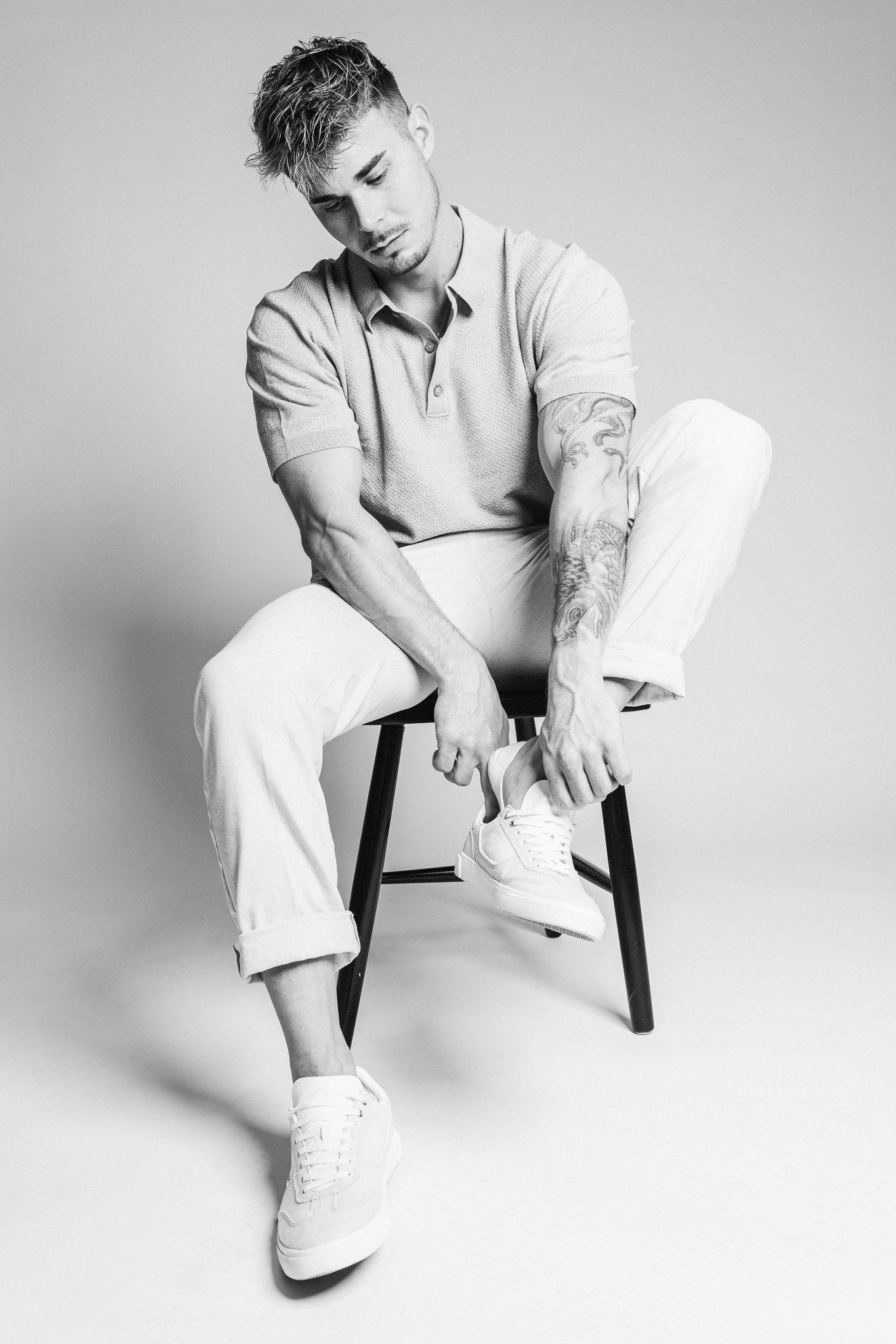 Stylish man in casual wear tying sneakers while sitting on a stool, embodying modern men's fashion with tattooed arms, a short-sleeved polo shirt, and rolled-up jeans in a monochrome aesthetic.