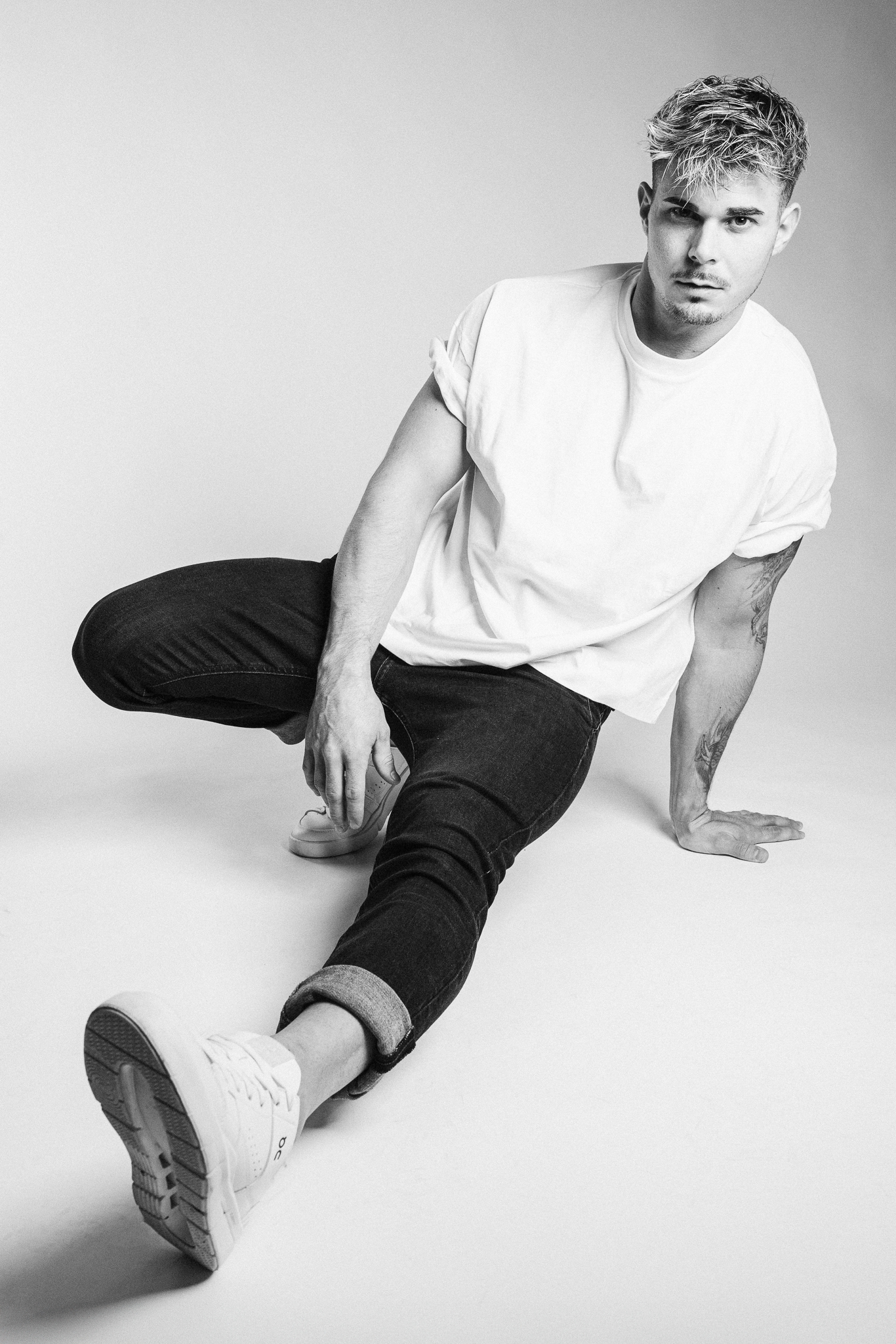 Black and white portrait of a stylish young man sitting on the floor, wearing a white t-shirt, dark jeans, and white sneakers, showcasing a casual fashion look.