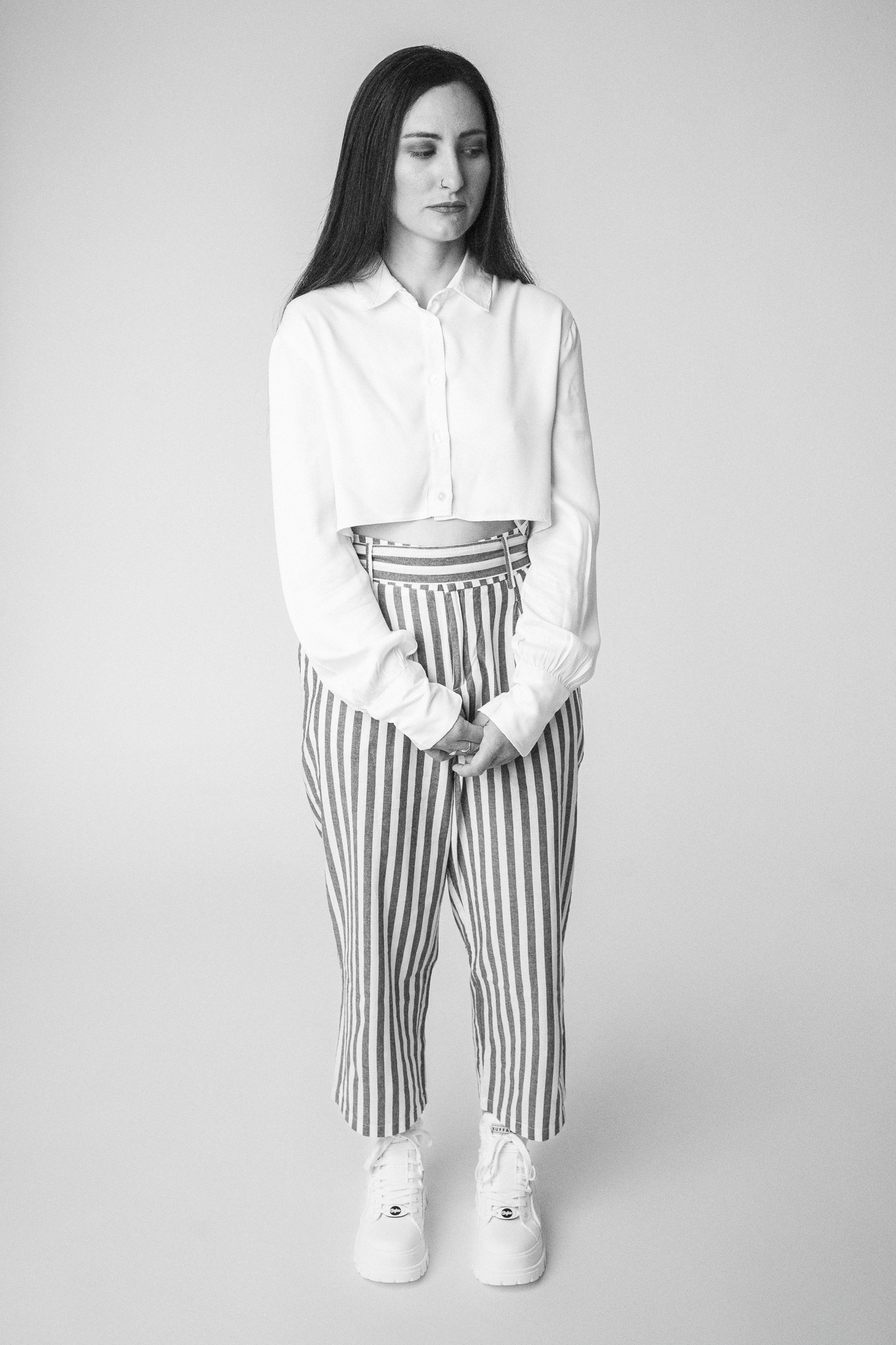 Grayscale photo of a woman wearing a cropped white shirt and striped high-waisted pants standing with hands clasped.