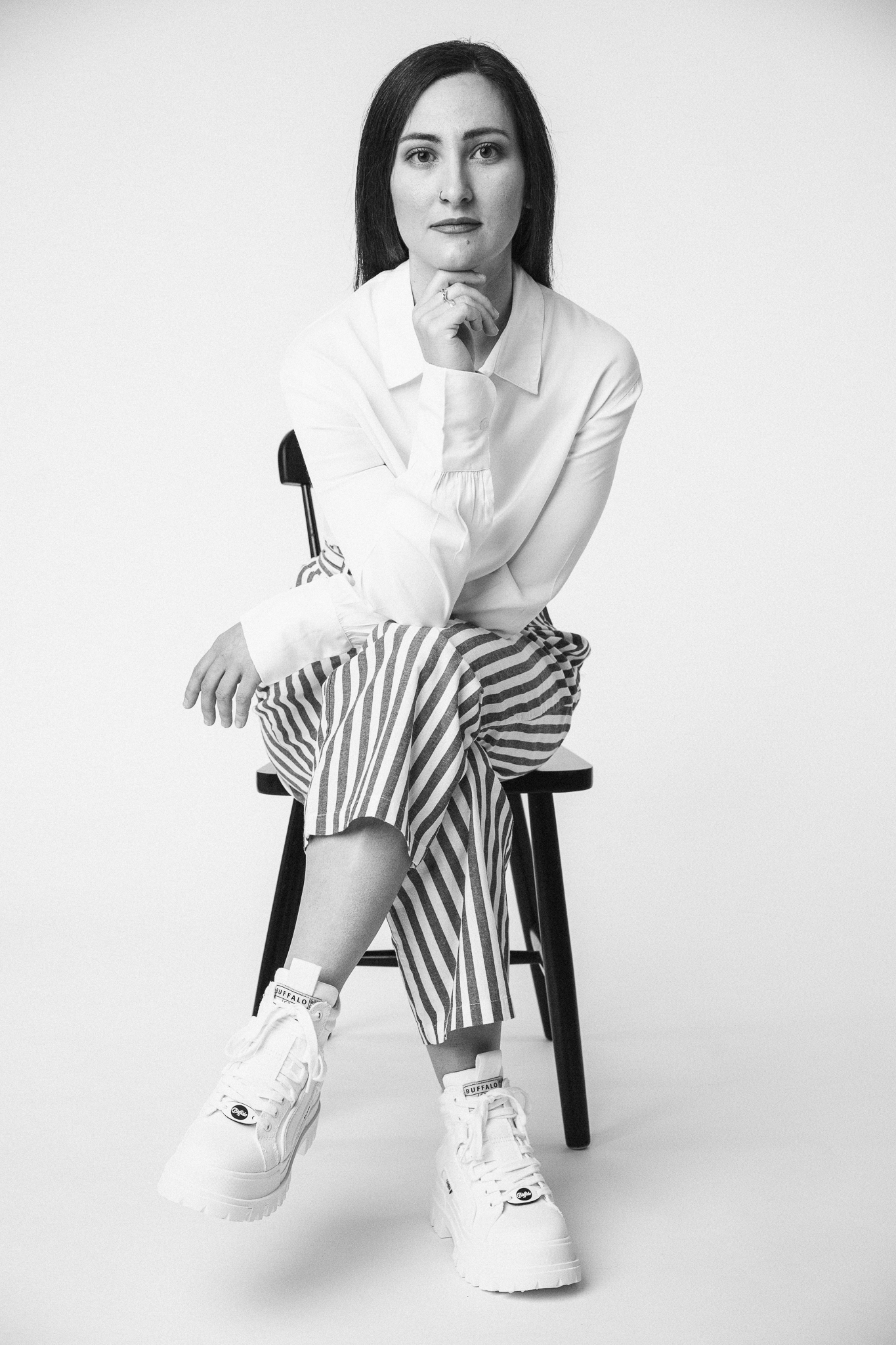 Black and white portrait of a woman sitting on a chair. She is wearing a white blouse, striped pants, and white sneakers, posing thoughtfully for a fashion photoshoot.