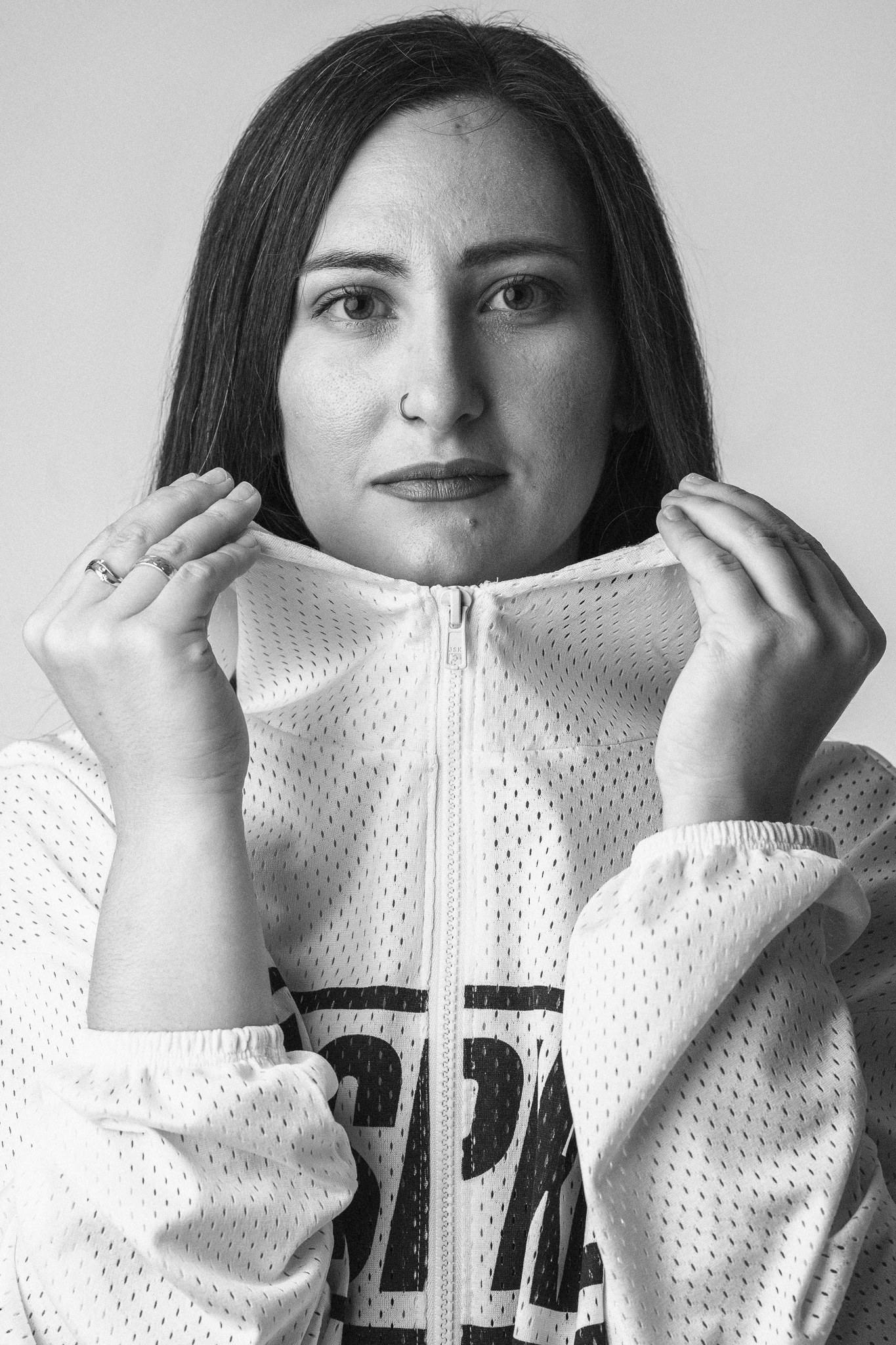 Black and white portrait of a woman in a zip-up jacket, holding the collar near her face. The jacket features a perforated pattern and lettering. The woman has a nose ring and an engaging, direct gaze. Perfect for articles on fashion, personal style, or monochromatic photography.