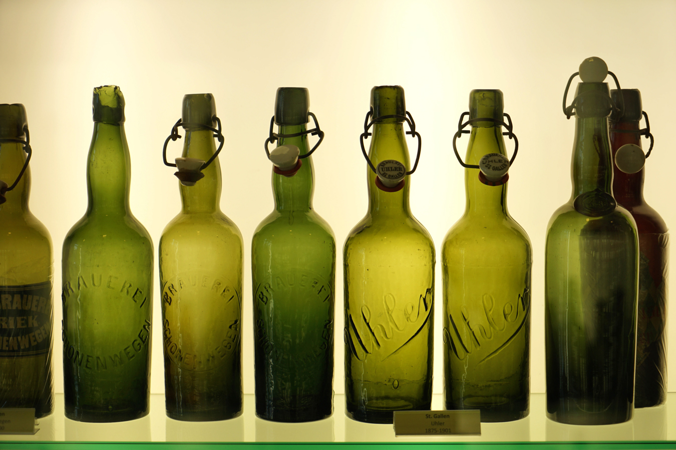Row of antique green glass bottles with ceramic stoppers against a lit background.