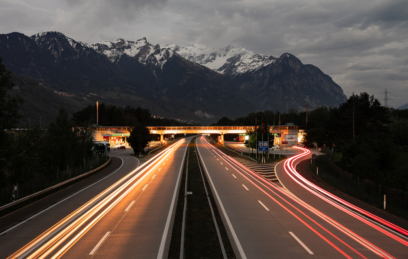 Highway at dusk with light trails from vehicles and snowy mountains in the background.