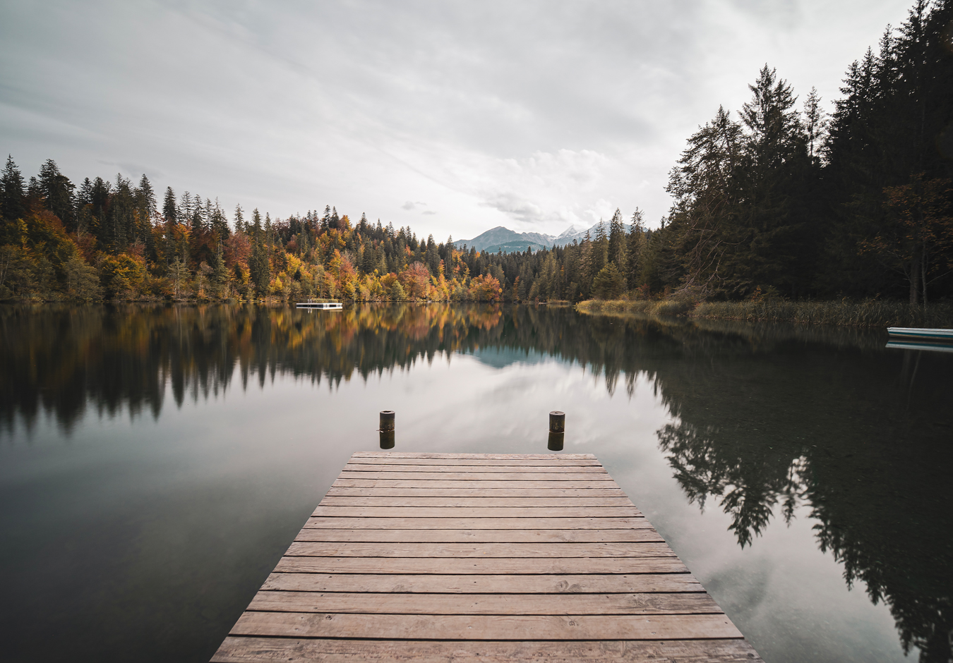 A serene lake view with a wooden dock, surrounded by autumn-colored trees and distant mountains under a cloudy sky, reflecting in the water.