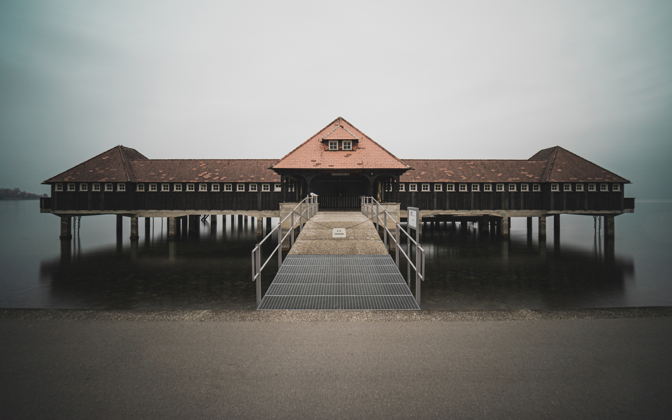 A wooden pavilion on stilts over calm water, featuring a peaked roof and a wide walkway leading to its entrance.