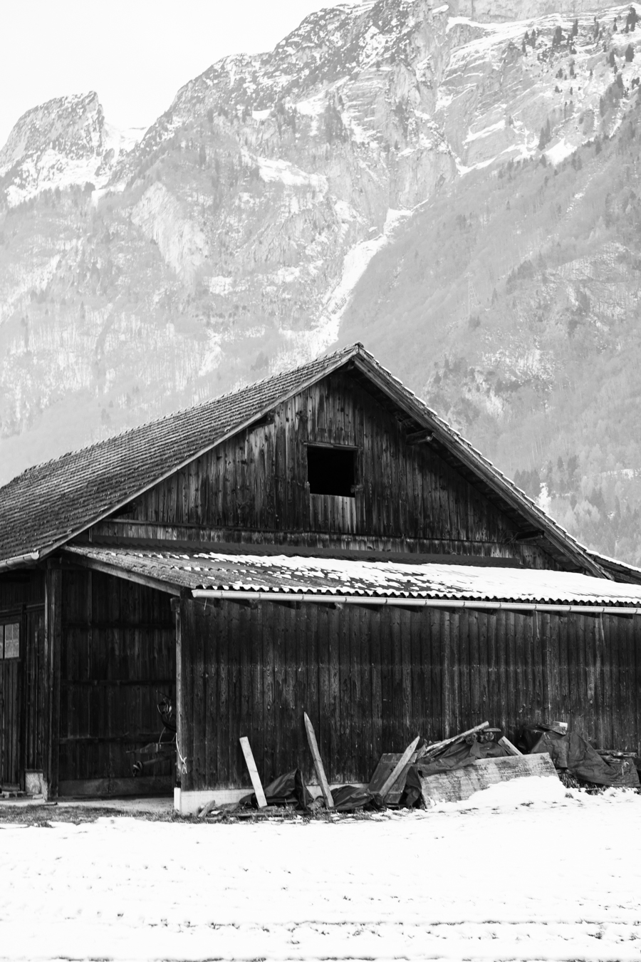 A rustic wooden barn sits against a backdrop of snow-capped mountains, captured in monochrome, evoking a serene winter landscape.