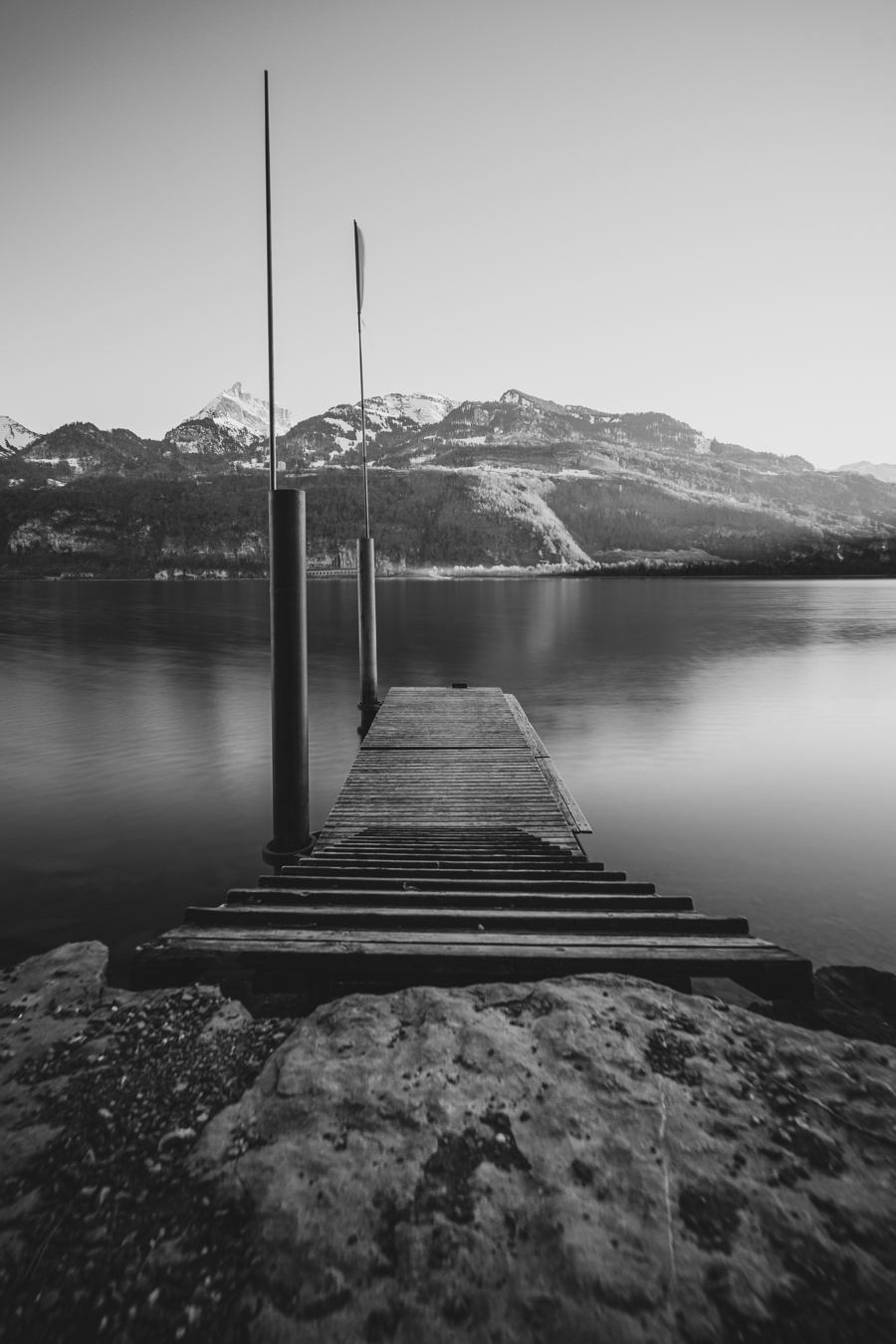 A serene black and white image of a wooden dock extending into still water, framed by mountains with snow-capped peaks in the background.