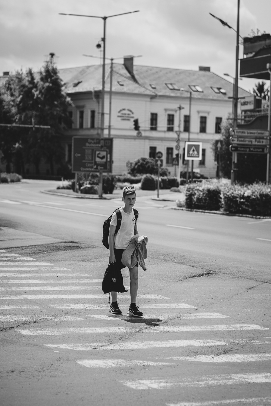 A young person walks across a zebra crossing, carrying a bag and a jacket, with buildings and traffic signs in the background.