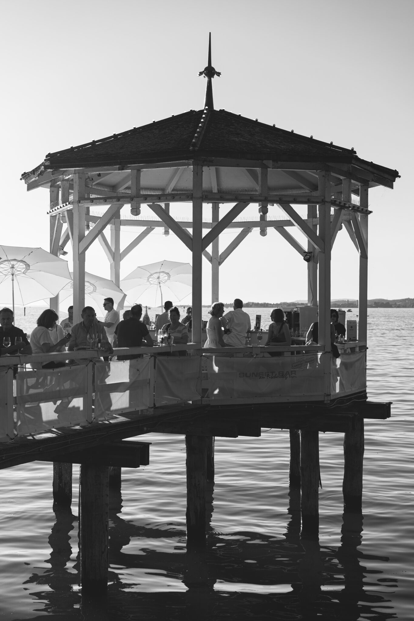 Black and white photo of people socializing under a lakeside gazebo. Umbrellas, warm atmosphere, reflections on water. Relaxed, scenic setting.