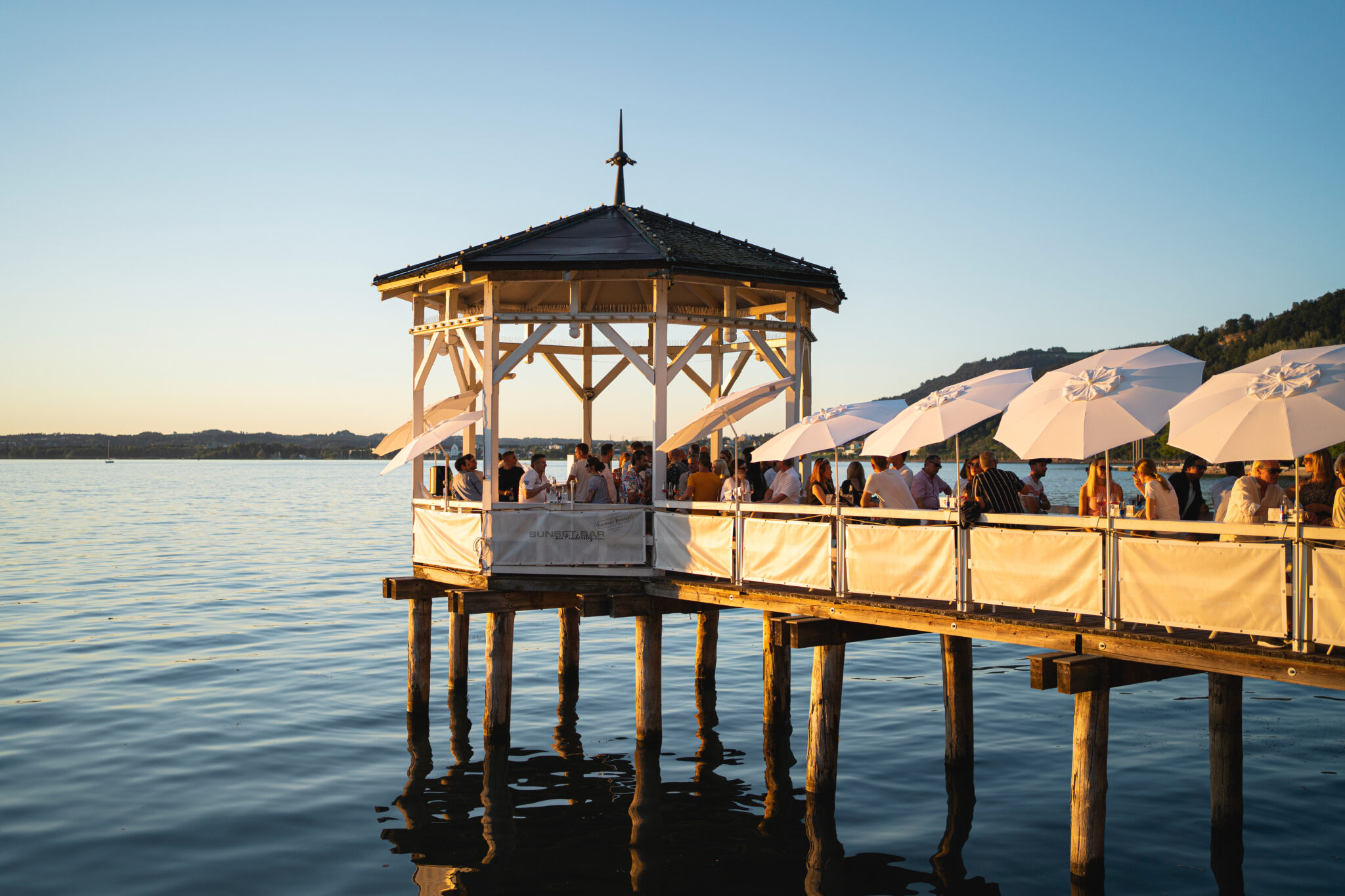 A serene lakeside scene at sunset with a gazebo on a wooden pier. People enjoy drinks under white umbrellas, casting soft reflections on the water.