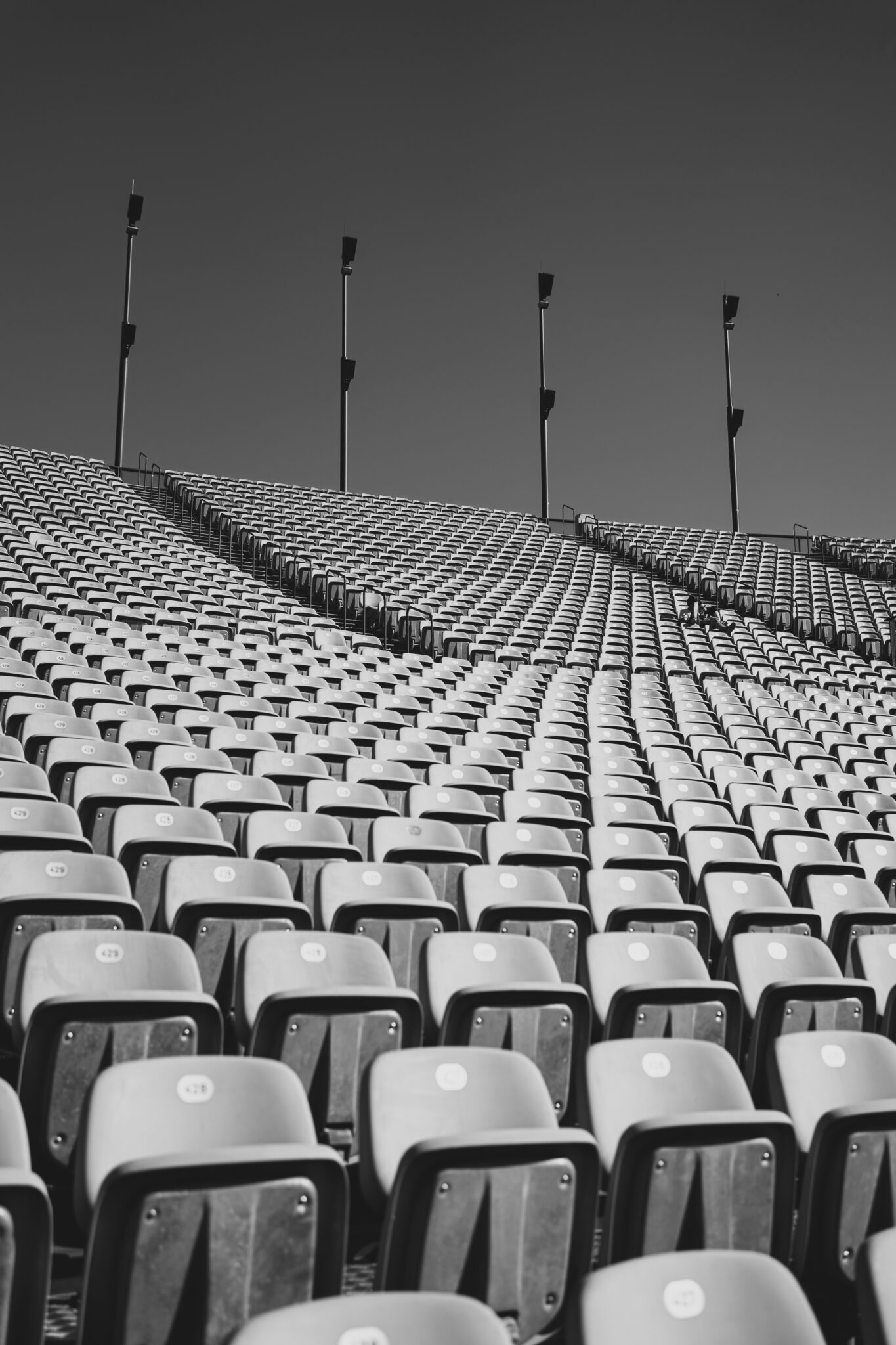 Black and white image of an empty stadium with rows of seats curving upwards. Tall floodlights stand overhead, creating a desolate, quiet atmosphere.