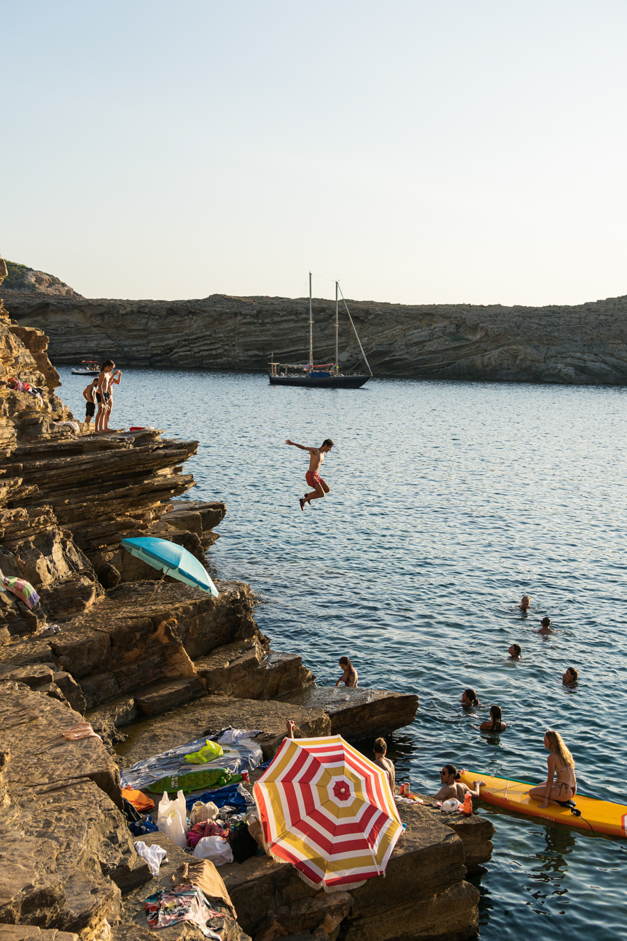 A sunlit rocky beach scene with swimmers in the water, a colorful umbrella, and a person jumping off a cliff. A sailboat is anchored nearby.