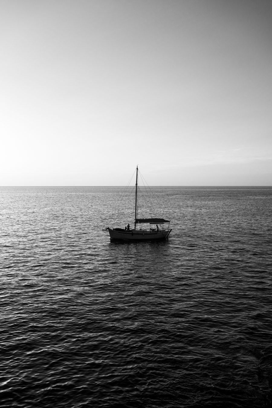 A solitary sailboat on calm waters, silhouetted against a serene, monochromatic sky at dusk. Two figures are visible on the boat.