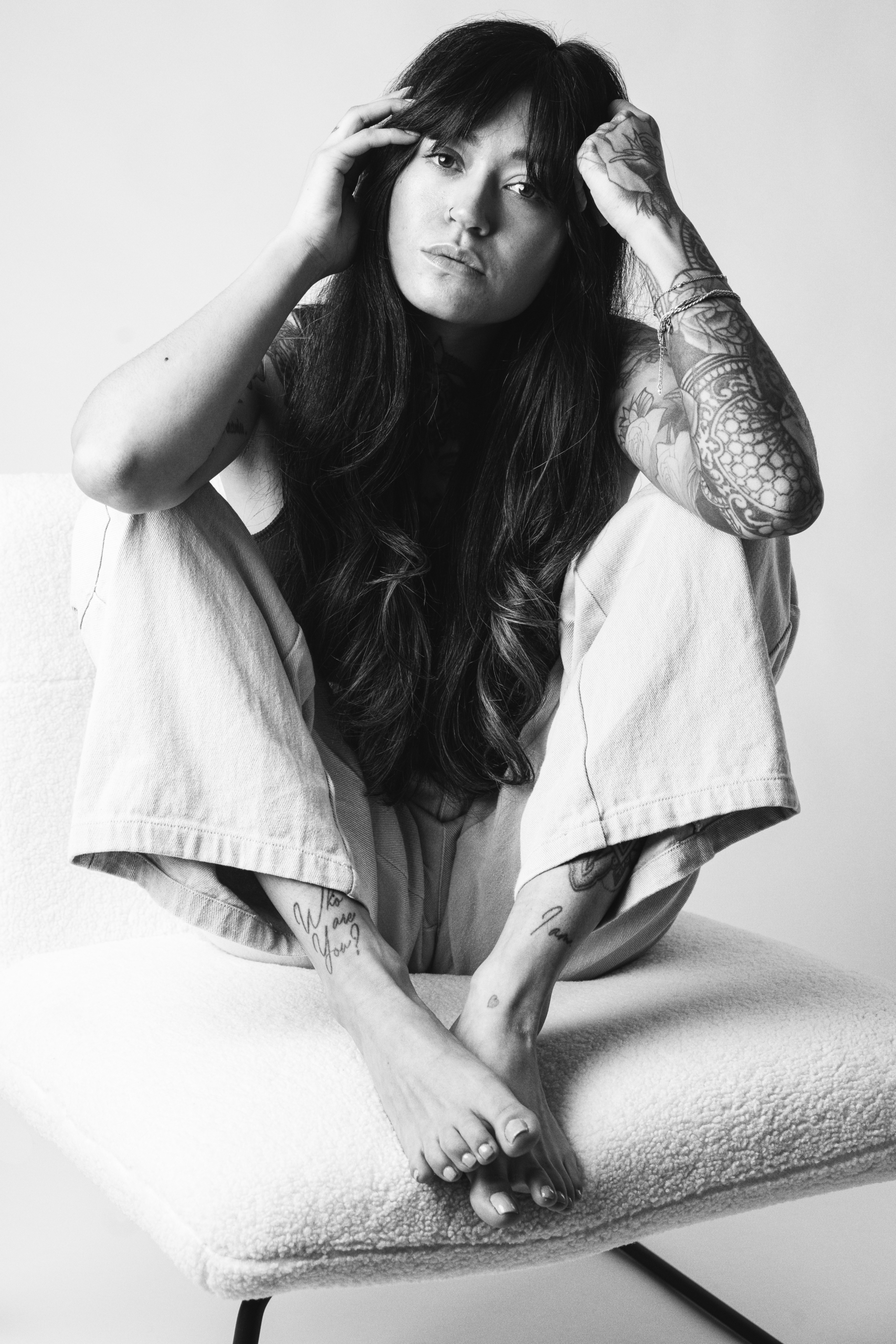 A person sits on a plush chair, hugging their knees with intricate tattoos visible on their arms and legs, set against a neutral background.