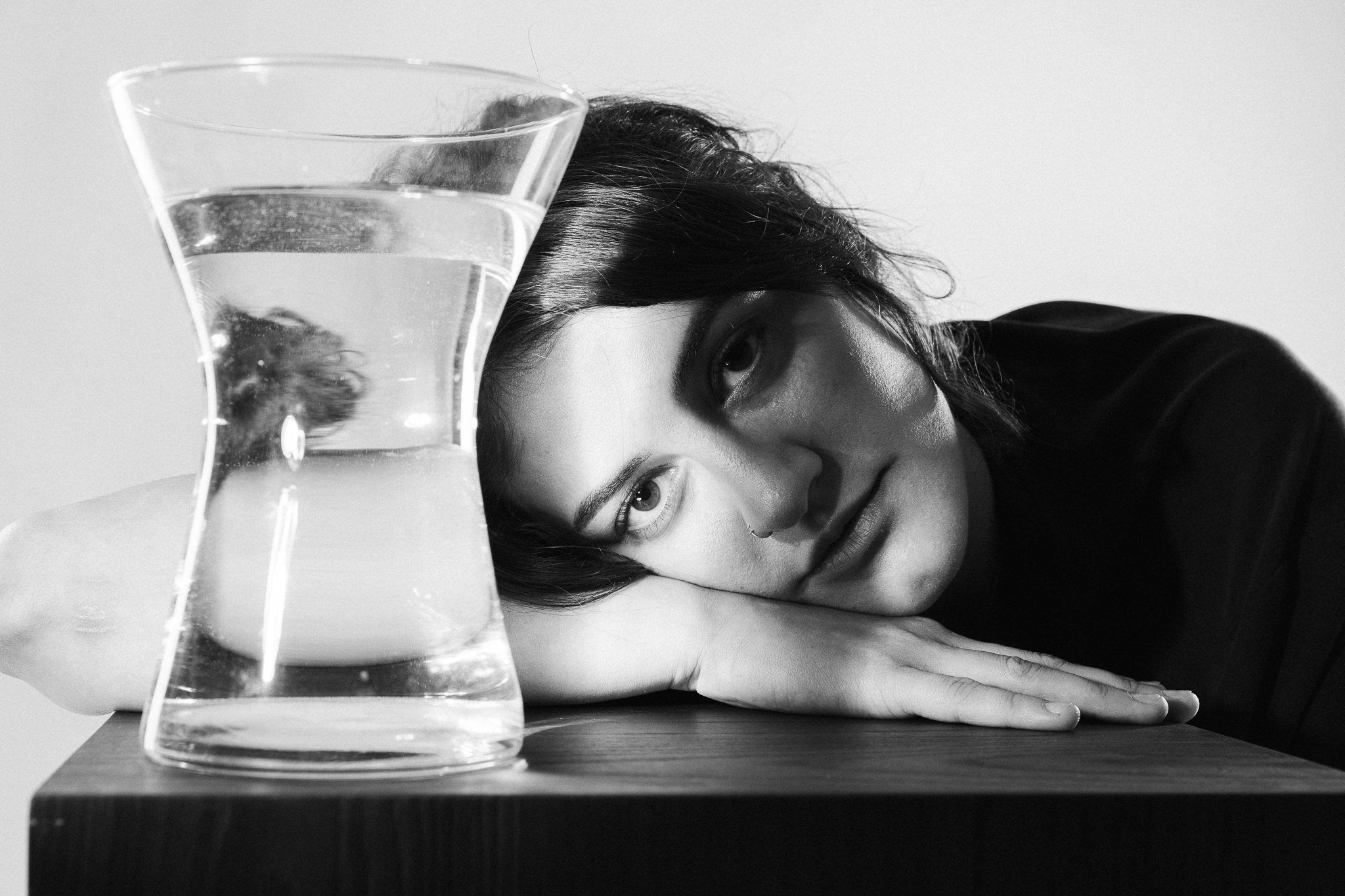 A woman rests her head on a table beside a glass of water, which distorts her reflection. The black-and-white image conveys a contemplative mood.