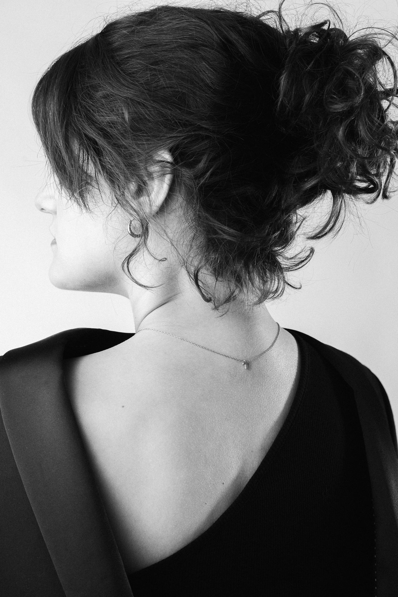 Black and white image of a woman viewed from the back. She has curly hair in a loose updo, wearing a one-shoulder top, exuding elegance and simplicity.